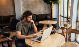 Ein Mann sitzt in einem Café an einem runden Holztisch und arbeitet konzentriert an einem Laptop. Neben ihm stehen eine Tasse Kaffee und ein Glas Wasser. Im Hintergrund sind weitere Tische und Stühle zu sehen.
